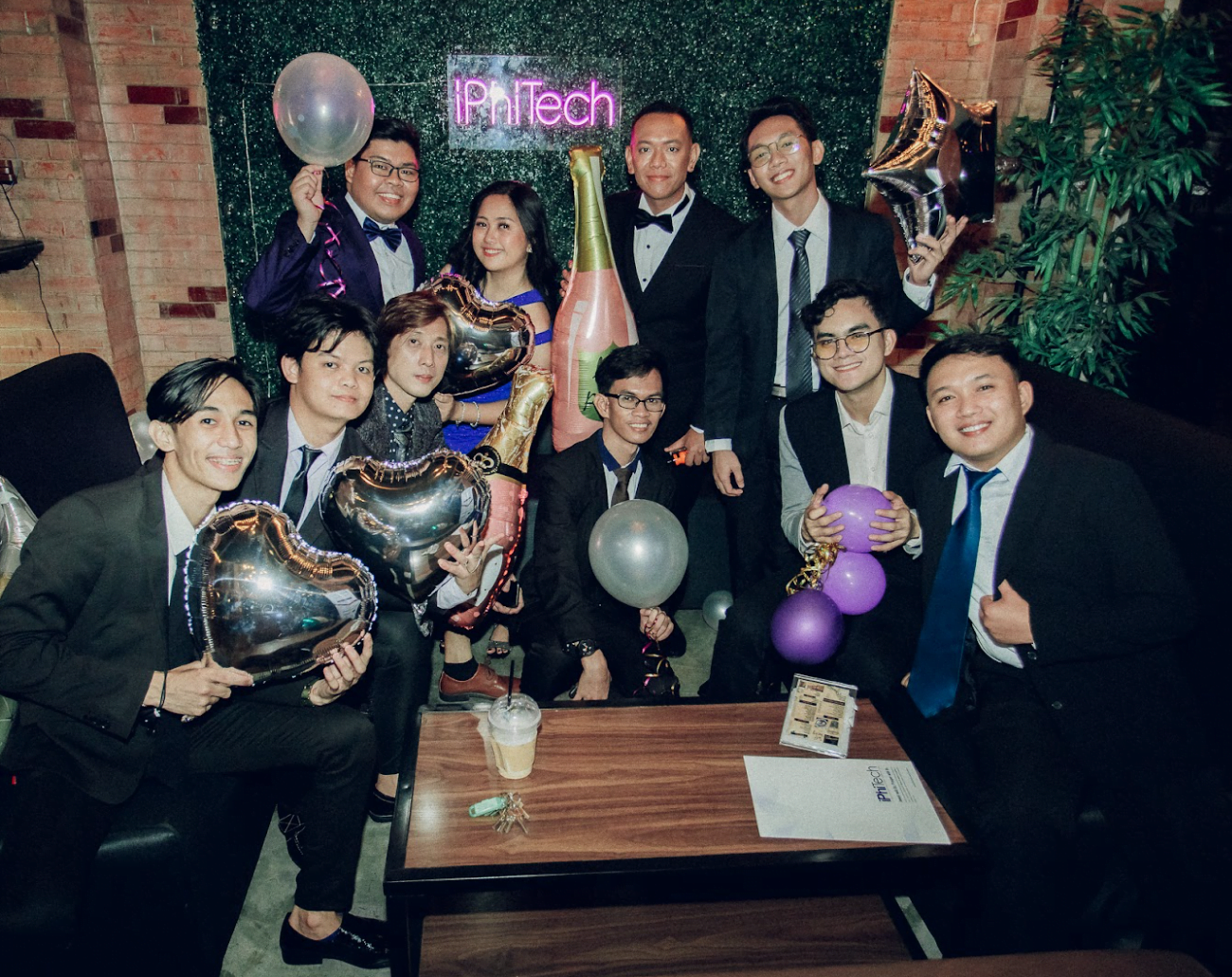 group of people having a party in a formal attire with balloons and a green backdrop with "iPhiTech" word